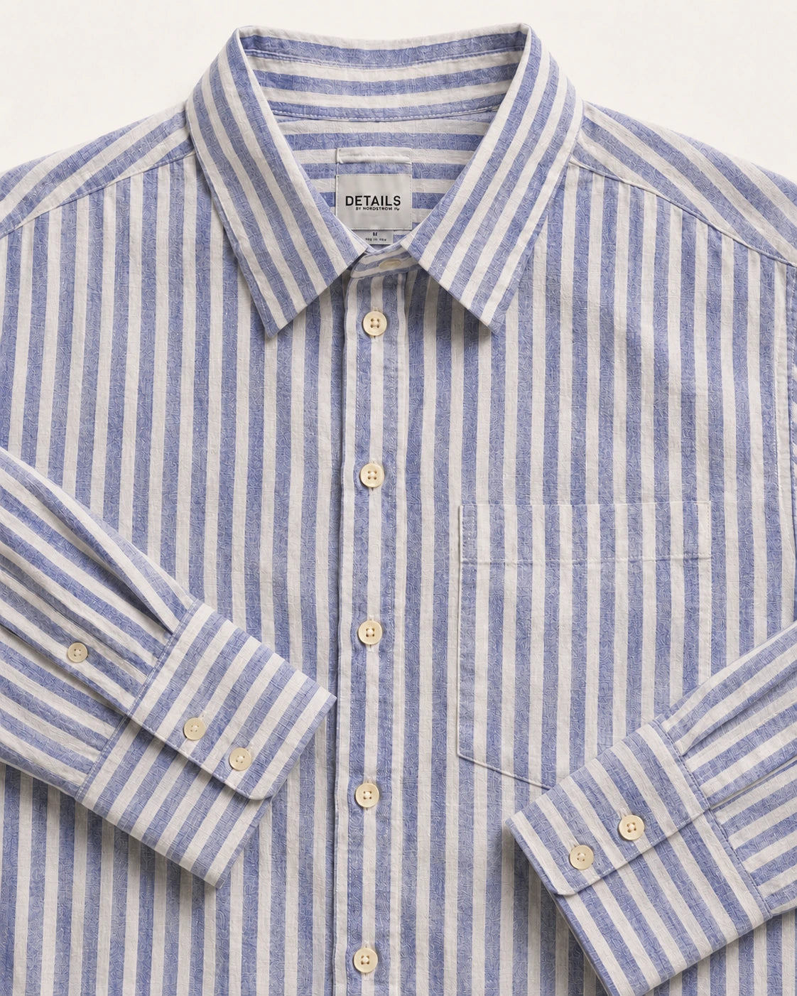 A close-up of a blue and white striped long-sleeve button-down shirt with a pointed collar, chest pocket, and light-colored buttons. The brand name 'DETAILS' is visible on the inner neck label.