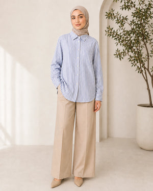 A woman wearing a light blue and white vertically striped button-up shirt, beige wide-leg trousers, and a beige hijab stands in a bright, modern room with a potted plant.