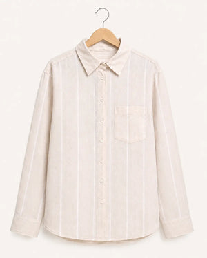 A light beige long-sleeve button-up shirt with thin white vertical stripes, featuring a pointed collar and a single chest pocket, hanging on a wooden hanger against a solid off-white background.
