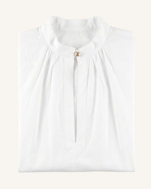 A folded white blouse with a high gathered collar and a small gold button at the neckline, shown on a plain light beige background.