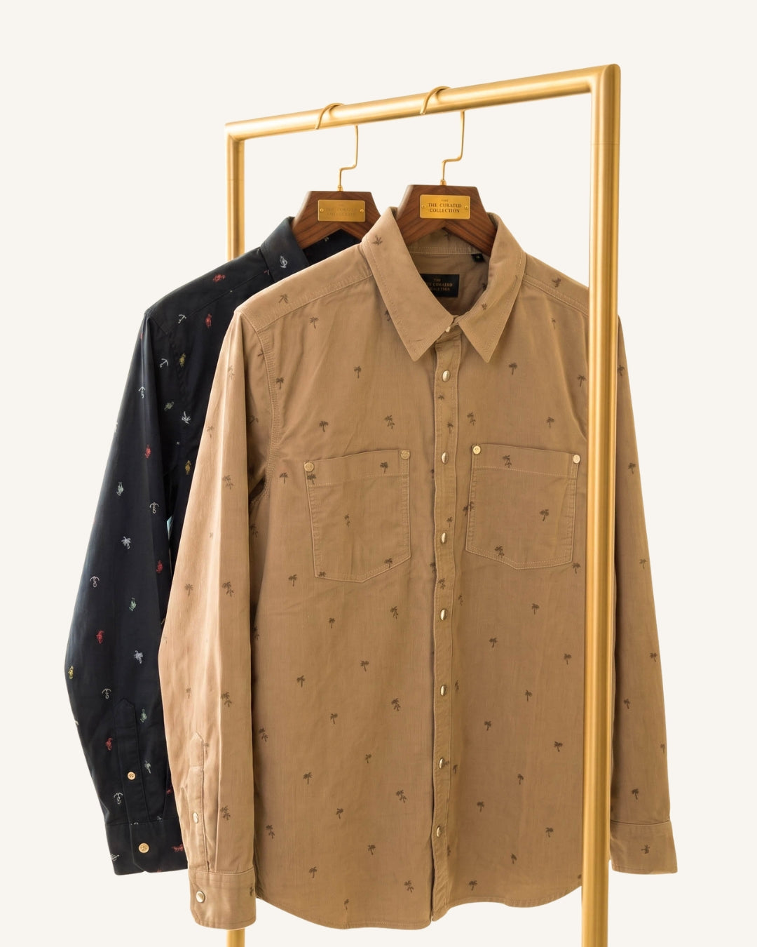 A tan long-sleeve button-up shirt with a small all-over embroidered pattern, hanging on a wooden hanger from a gold clothing rack, with a navy blue patterned shirt behind it.