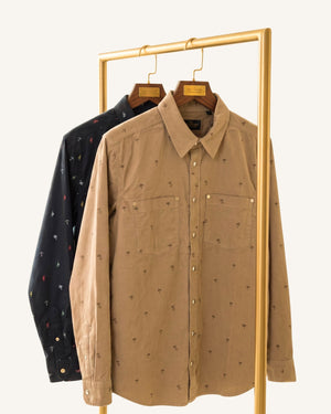 A tan long-sleeve button-up shirt with a small all-over embroidered pattern, hanging on a wooden hanger from a gold clothing rack, with a navy blue patterned shirt behind it.