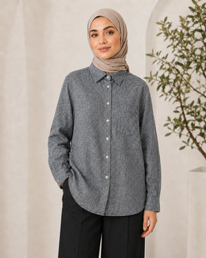 A woman wearing a gray long-sleeve button-up shirt with a subtle textured pattern, paired with black trousers and a beige hijab, standing against a neutral cream-colored background with a plant on the right.