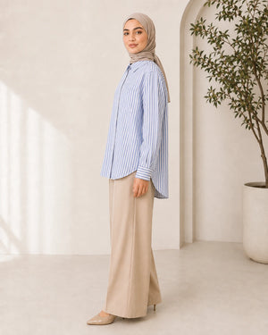 A woman wearing a light blue and white vertically striped button-up shirt, beige wide-leg trousers, and a beige hijab stands in profile in a bright, modern room with a potted plant.
