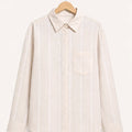 A light beige long-sleeve button-up shirt with thin white vertical stripes, featuring a pointed collar and a single chest pocket, hanging on a wooden hanger against a solid off-white background.