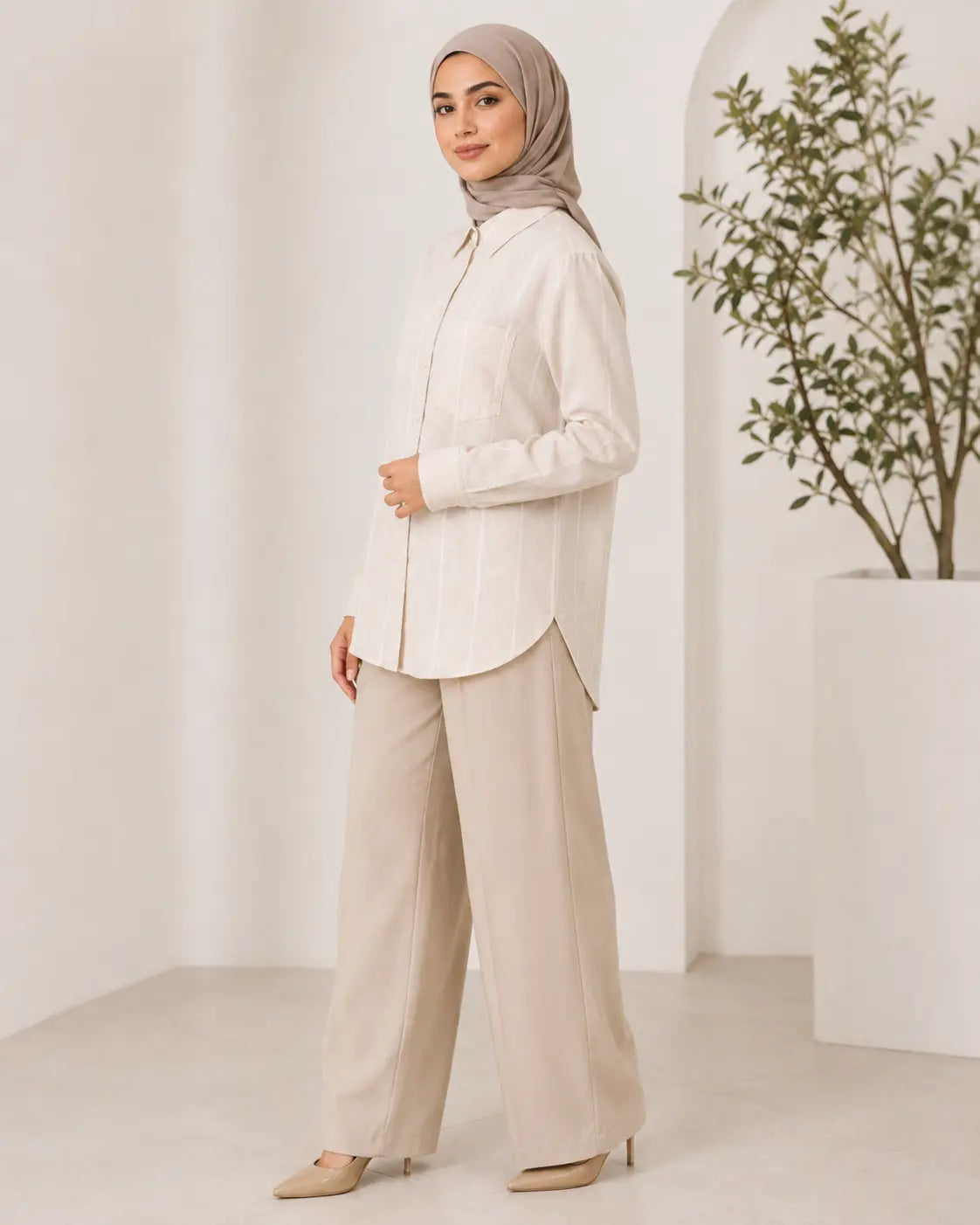 A woman wearing a light beige/ivory long-sleeve button-up shirt, wide-leg beige trousers, and a taupe hijab, standing in a bright, minimalist interior with a potted plant.