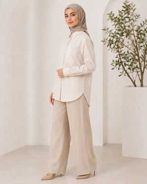 A woman wearing a light beige/ivory long-sleeve button-up shirt, wide-leg beige trousers, and a taupe hijab, standing in a bright, minimalist interior with a potted plant.