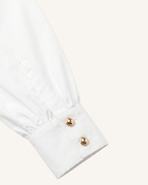 Close-up of a white dress shirt cuff with two gold-tone buttons on a light cream background.