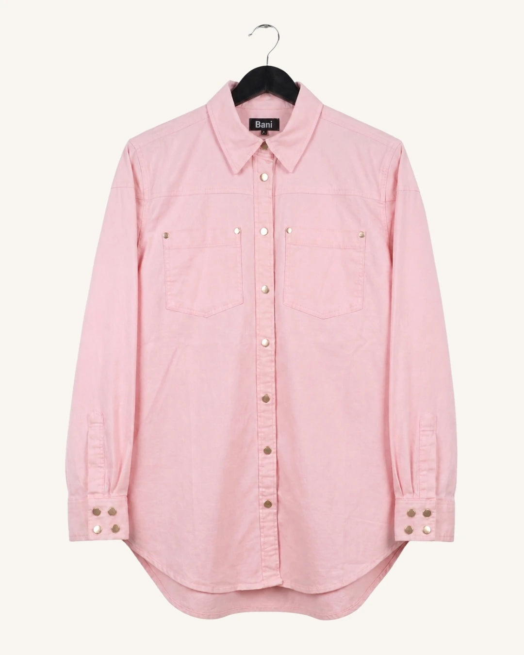 A light pink long-sleeve button-up shirt with two chest pockets and decorative metal snap details, hanging on a black hanger against a plain off-white background.