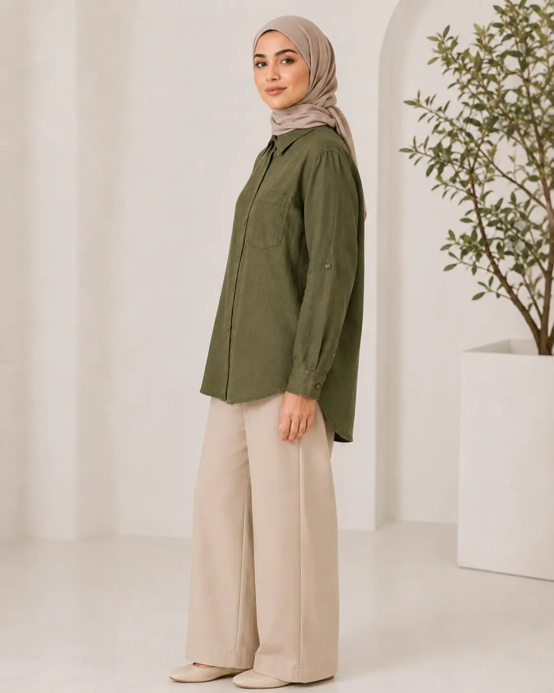 A woman wearing an olive green button-up shirt, beige wide-leg trousers, and a taupe hijab stands in profile against a bright, minimalist white studio background with a potted plant.