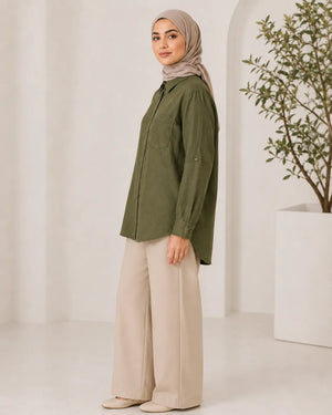 A woman wearing an olive green button-up shirt, beige wide-leg trousers, and a taupe hijab stands in profile against a bright, minimalist white studio background with a potted plant.