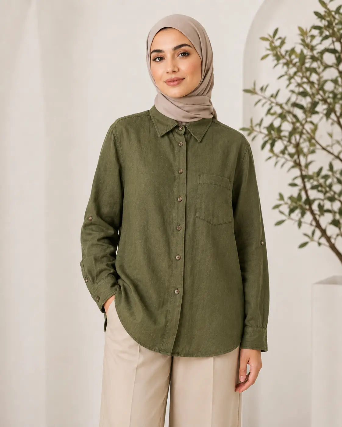 A woman wearing a muted olive green long-sleeve button-up shirt with a chest pocket, paired with beige trousers and a taupe hijab, standing in a bright indoor setting with a white wall and a plant in the background.