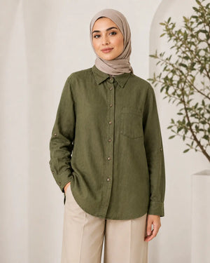 A woman wearing a muted olive green long-sleeve button-up shirt with a chest pocket, paired with beige trousers and a taupe hijab, standing in a bright indoor setting with a white wall and a plant in the background.