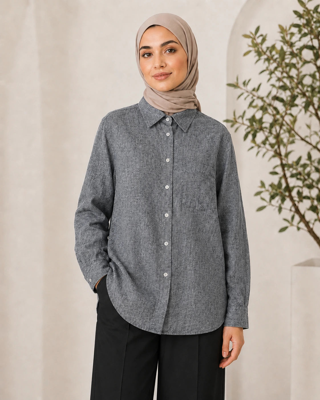 A woman wearing a gray long-sleeve button-up shirt with a subtle textured pattern, paired with black trousers and a beige hijab, standing against a neutral cream-colored background with a plant on the right.