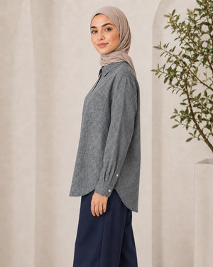 A woman wearing a heather gray long-sleeve button-up shirt and navy wide-leg pants, paired with a beige hijab, standing in a neutral indoor setting with a potted plant.