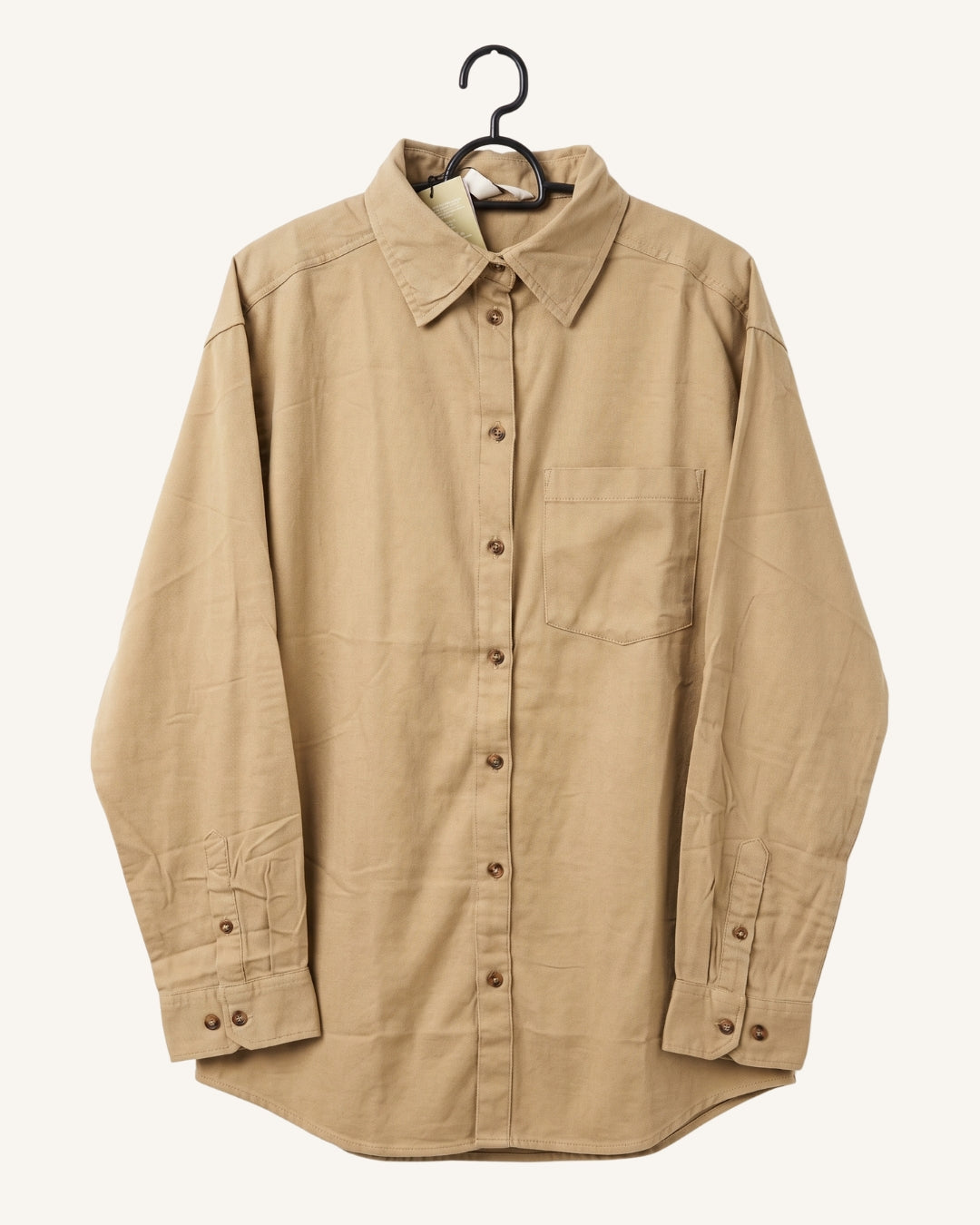 A beige long-sleeve button-up shirt with a pointed collar and a single chest pocket, hanging on a black hanger against a light cream background.